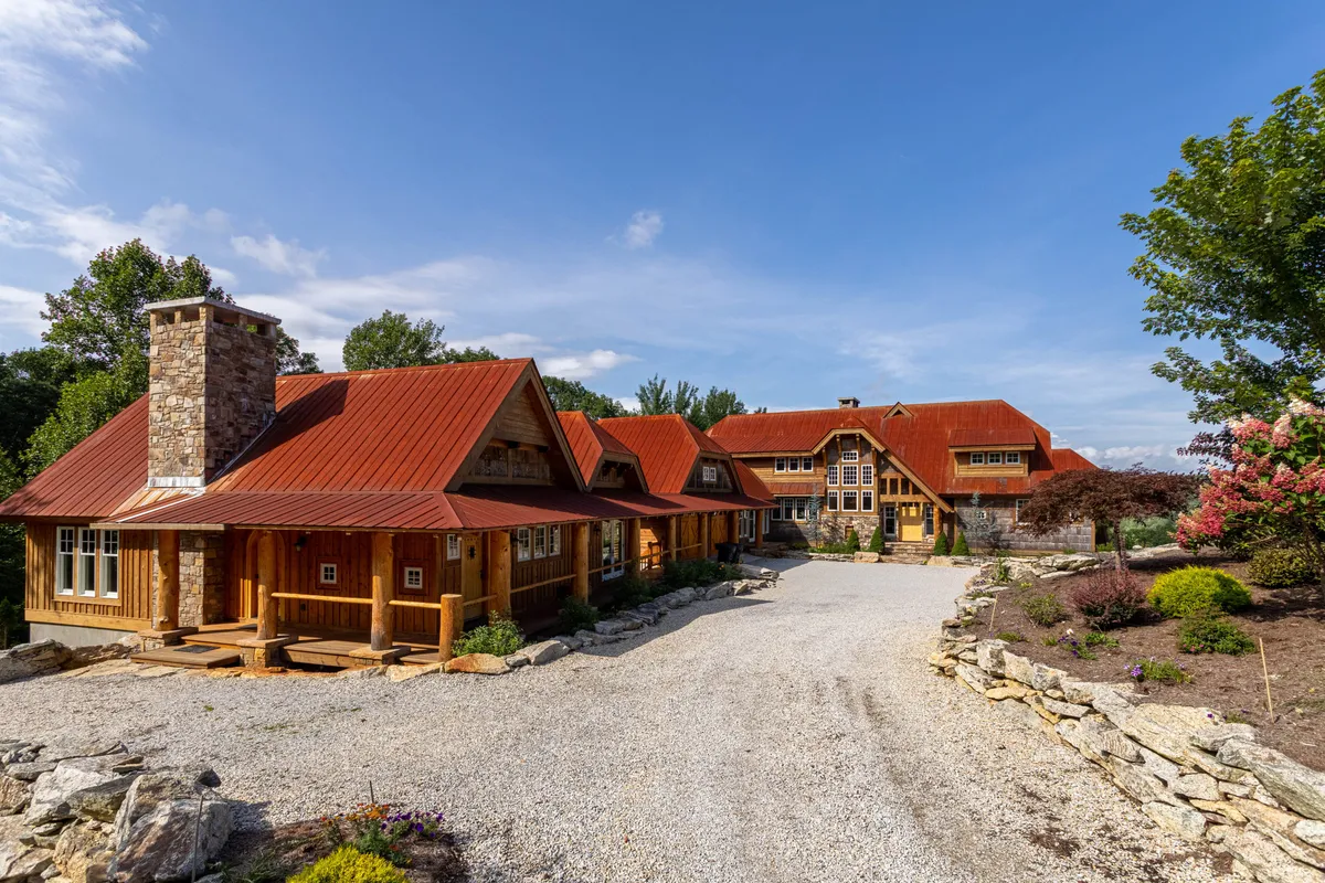 The image showcases the exterior of a welcoming wooden lodge with distinct red metal roofs, a tall stone chimney, and beautifully landscaped gardens surrounding the property. The setting suggests an ideal location for family gatherings and outdoor activities, with a peaceful, nature-rich backdrop.