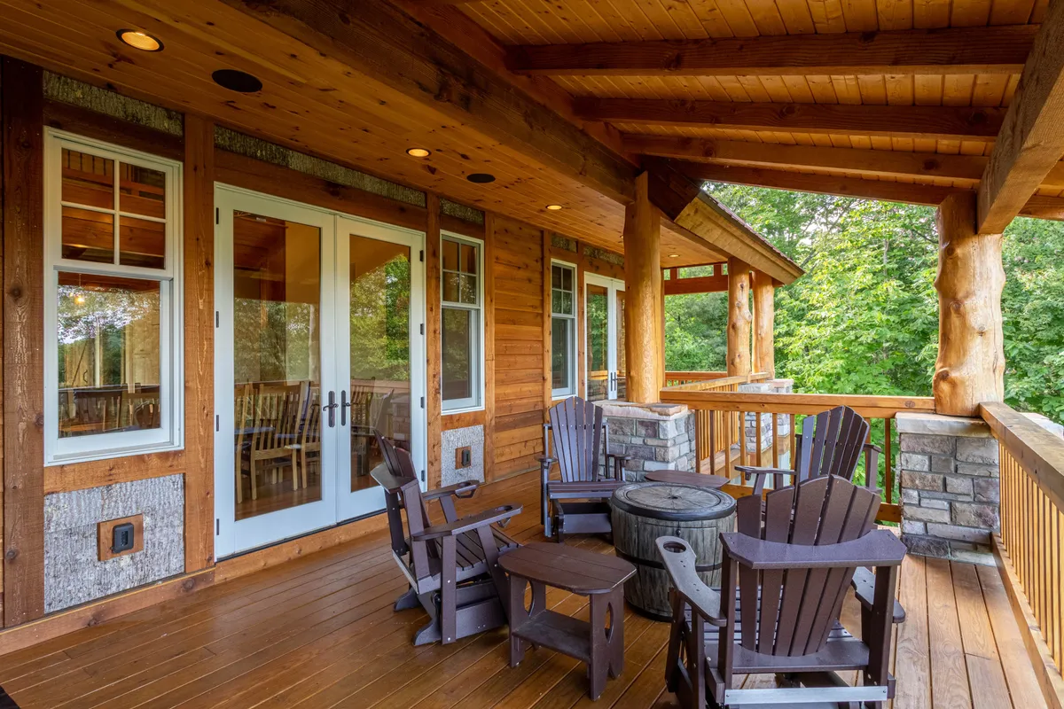 A cozy wooden deck is outfitted with rocking chairs and a barrel table, surrounded by lush greenery and large windows reflecting sunset hues. The space invites relaxation, socializing, and enjoying the outdoors—perfect for families looking to unwind after a day of adventure.