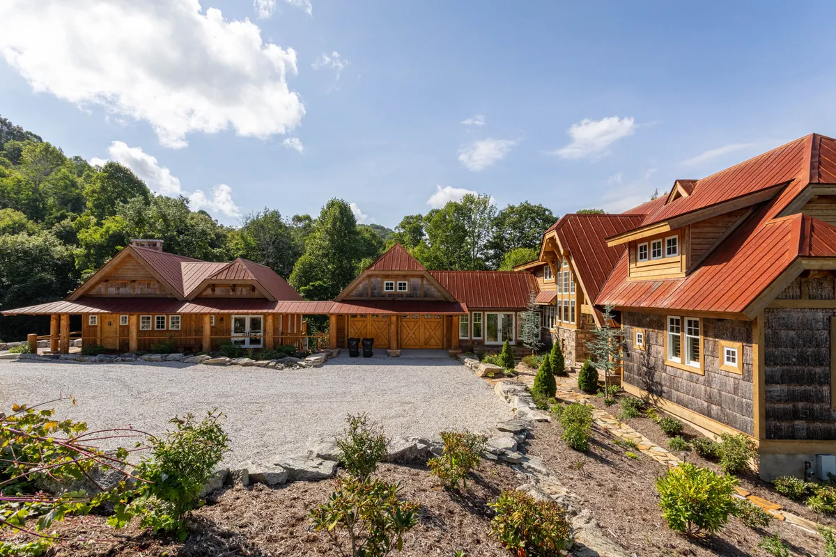 A charming exterior view of rustic wooden buildings with red metal roofs, a gravel driveway, and lush greenery all around. The overall scene offers a welcoming sense of arrival, ideal for family or group retreats in a secluded natural setting.