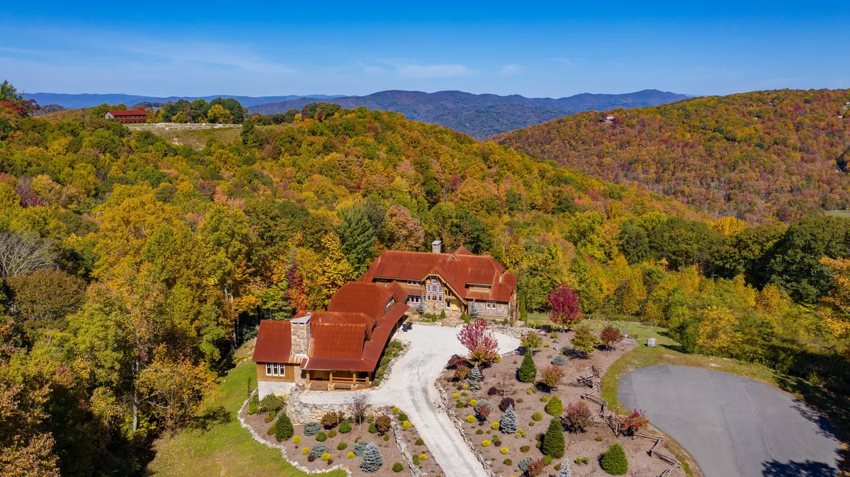 The photo features the main house with a vibrant red roof nestled among colorful foliage and meticulous gardens, set against a backdrop of mountains. The inviting outdoor areas suggest opportunities for hiking, autumn festivals, or simply relaxing on the porch.