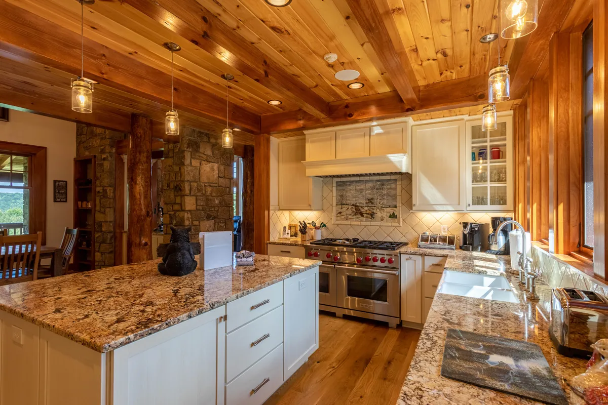 A spacious kitchen featuring a central island with granite countertops, stainless steel appliances, and a striking wooden ceiling accented by a stone wall. The kitchen exudes warmth and modern comfort, inviting guests to cook, gather, and enjoy meals together.