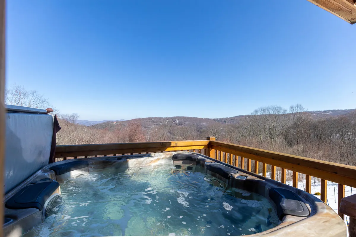 This exterior image captures a bubbling hot tub on a wooden deck with a panoramic view of the surrounding mountains. The wooden railing frames the scene, which feels serene and private—an ideal spot for relaxation or stargazing.