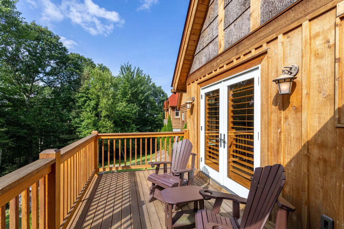 This image presents a welcoming wooden deck furnished with Adirondack chairs, framed by surrounding trees and double doors leading into the home. The setting exudes tranquility and invites guests to unwind while enjoying the outdoor ambiance.