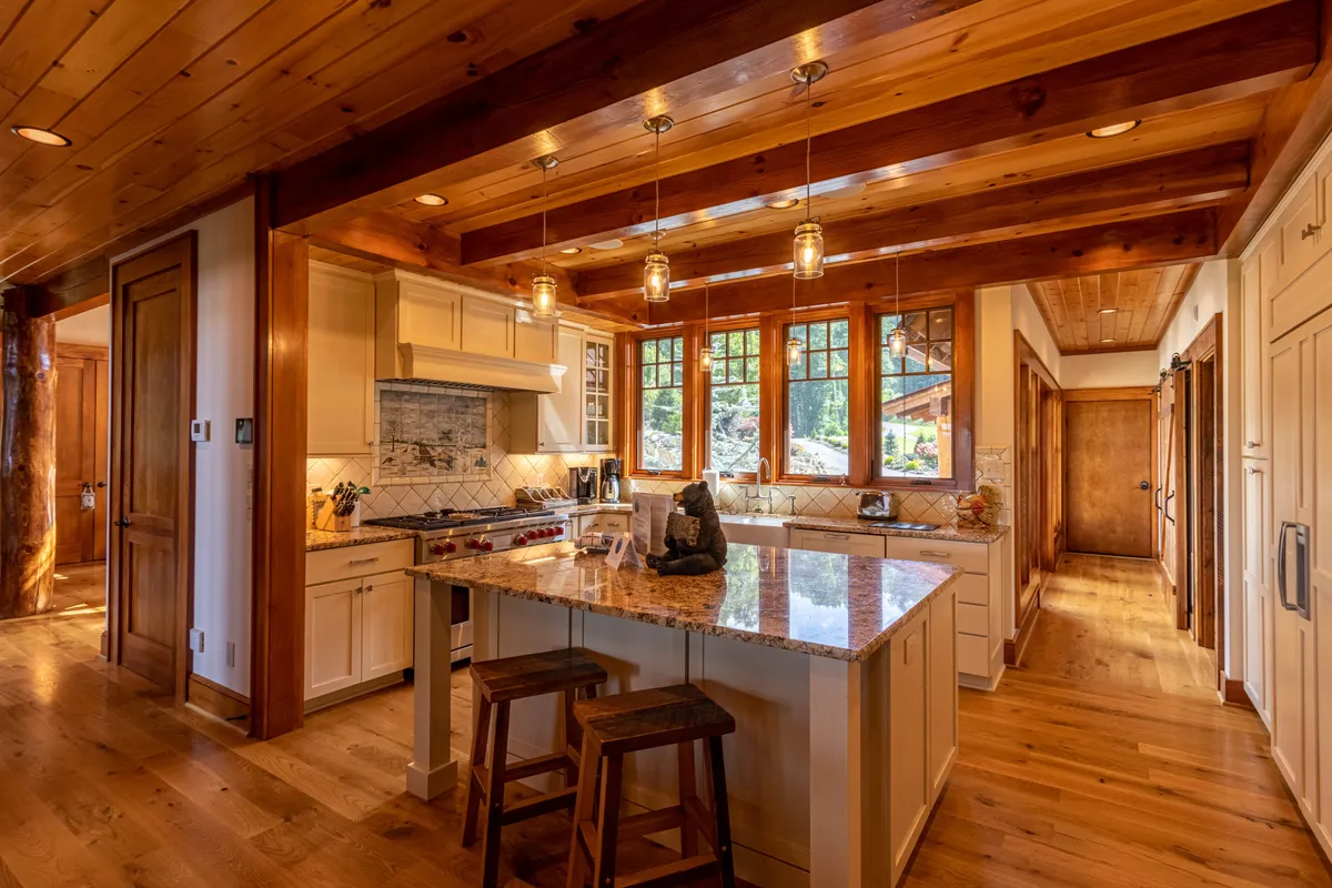 A chef-inspired kitchen featuring a granite countertop, wooden beams, a substantial kitchen island, and windows that offer glimpses of the outdoors. The kitchen is arranged for both entertaining and creating gourmet meals.