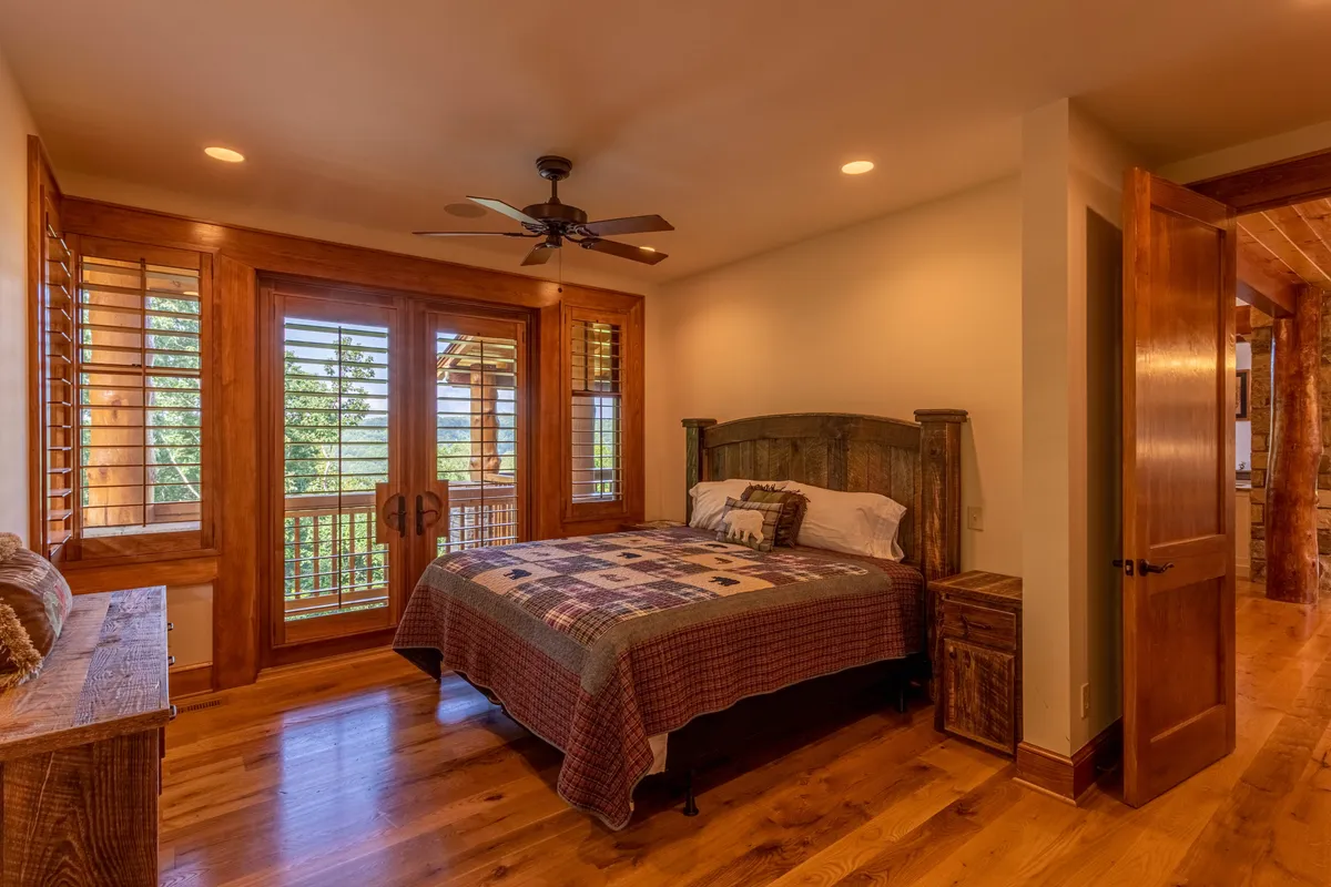A serene bedroom featuring a comfortable bed with a quilted blanket, wooden furniture, and large windows offering an expansive view. The space is bathed in natural light, highlighting the tranquil and cozy atmosphere perfect for relaxation.