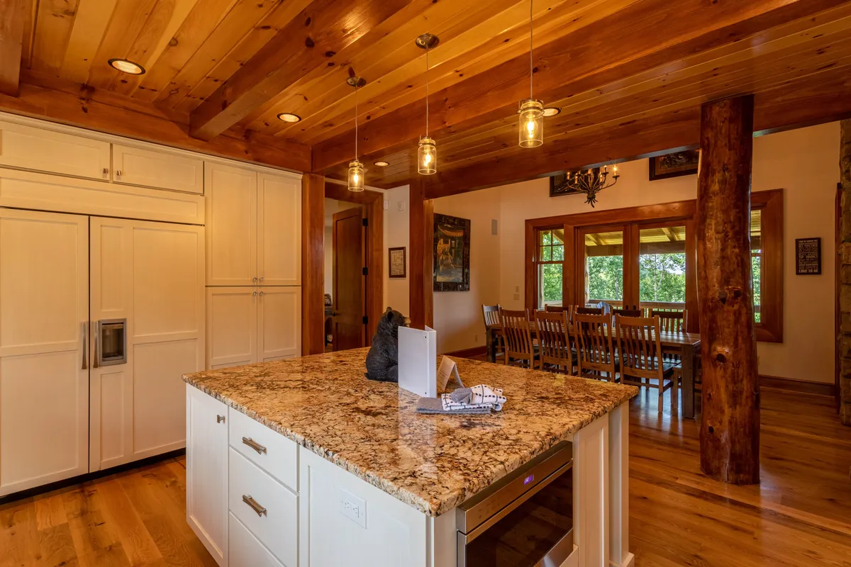 A beautifully designed kitchen and dining area featuring a substantial granite island, rustic wooden beams overhead, and an adjacent dining table with seating for a group. The open layout encourages connection between the kitchen and communal dining spaces, ideal for shared meals or conversations over coffee.