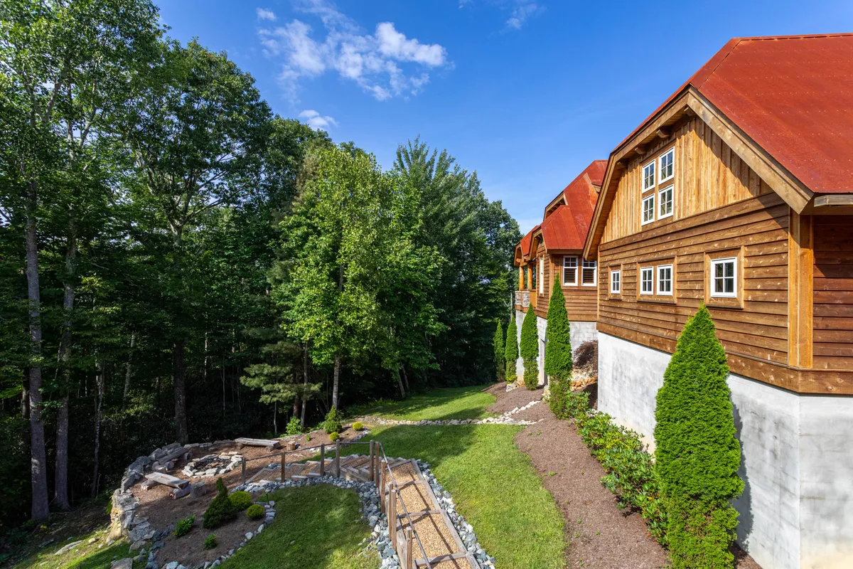 This exterior image presents a landscaped pathway leading to a wooden gathering area, surrounded by lush greenery and characteristic cabin architecture. It evokes feelings of tranquility and warmth, suggesting outdoor spaces ideal for gathering around a fire pit, sharing stories, and stargazing.