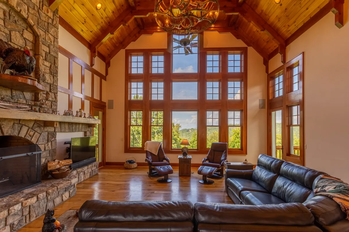 A spacious living room with large windows offering expansive mountain views, a striking stone fireplace as the focal point, a chandelier overhead, and comfortable leather seating including a sofa and recliners. Sunlight pours in, highlighting the room's cozy yet elegant ambiance—perfect for socializing, relaxing, or enjoying the scenery.