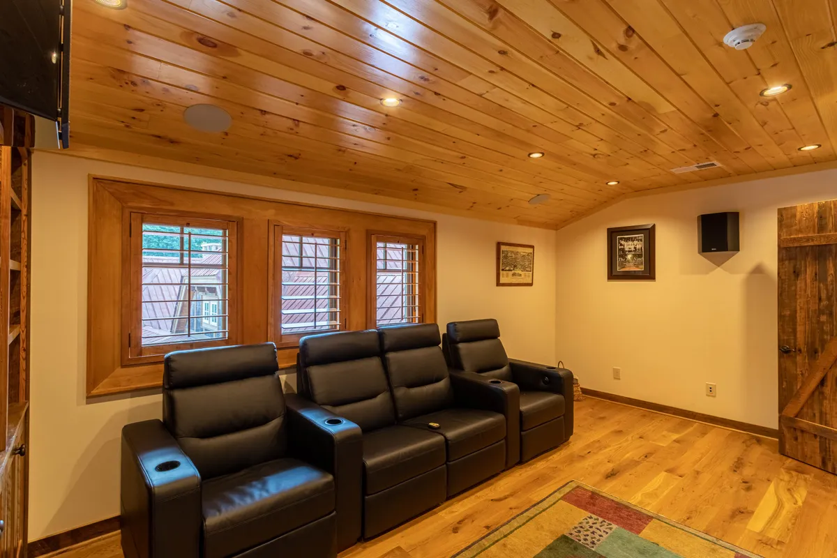 This interior image showcases a cozy movie room with four plush reclining theater seats arranged for an immersive viewing experience. The space features a wooden ceiling, windows with shutters for light control, and decorative wall art, creating a comfortable and intimate setting ideal for movie nights and gatherings.