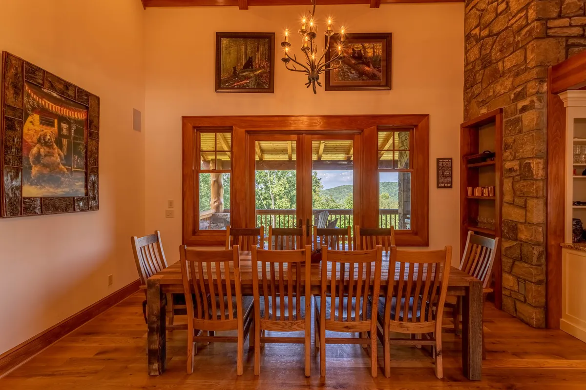 A warm, inviting dining space featuring a large wooden table set for ten, illuminated by a chandelier, and opening to a balcony through double doors. The stone wall and cozy lighting set the tone for memorable holiday celebrations or intimate gatherings with a view.