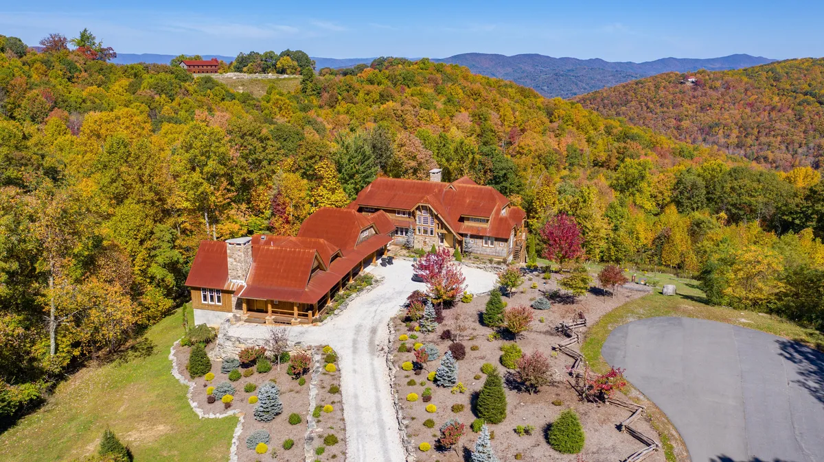 A broad exterior view of the main house with its distinctive red roof, surrounded by beautifully landscaped gardens awash with colorful foliage, set against a majestic mountain backdrop. The scene conveys tranquility and the allure of the mountain setting.