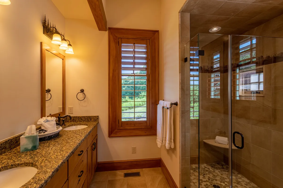 A spa-like bathroom featuring a double sink granite vanity, a large glass shower, and a window with scenic views. The abundance of morning sunlight creates a refreshing, high-end atmosphere.